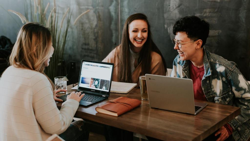 Drei Frauen sitzen lachend um einem Tisch mit ihren Laptops