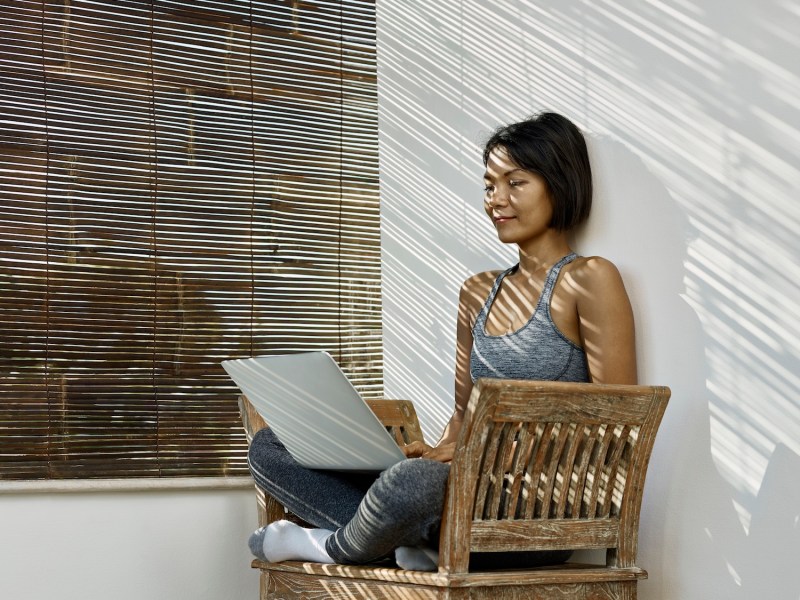 Eine Frau sitzt mit Laptop auf einer Bank im Schatten.