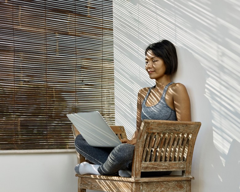 Eine Frau sitzt mit Laptop auf einer Bank im Schatten.