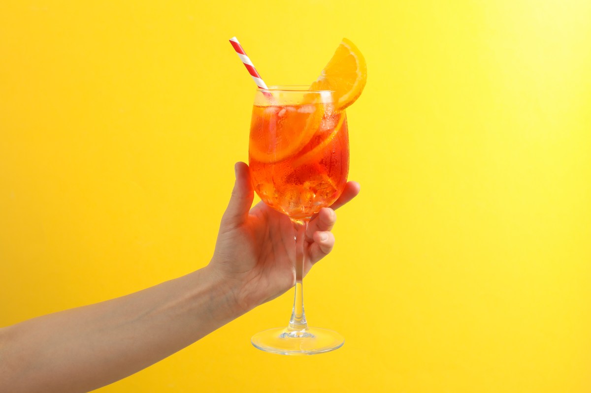 Aperol Spritz im Glas mit Strohhalm in der Hand auf gelbem Hintergrund