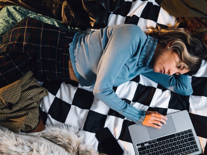 Eine junge Frau liegt auf dem Bett und arbeitet am Laptop.