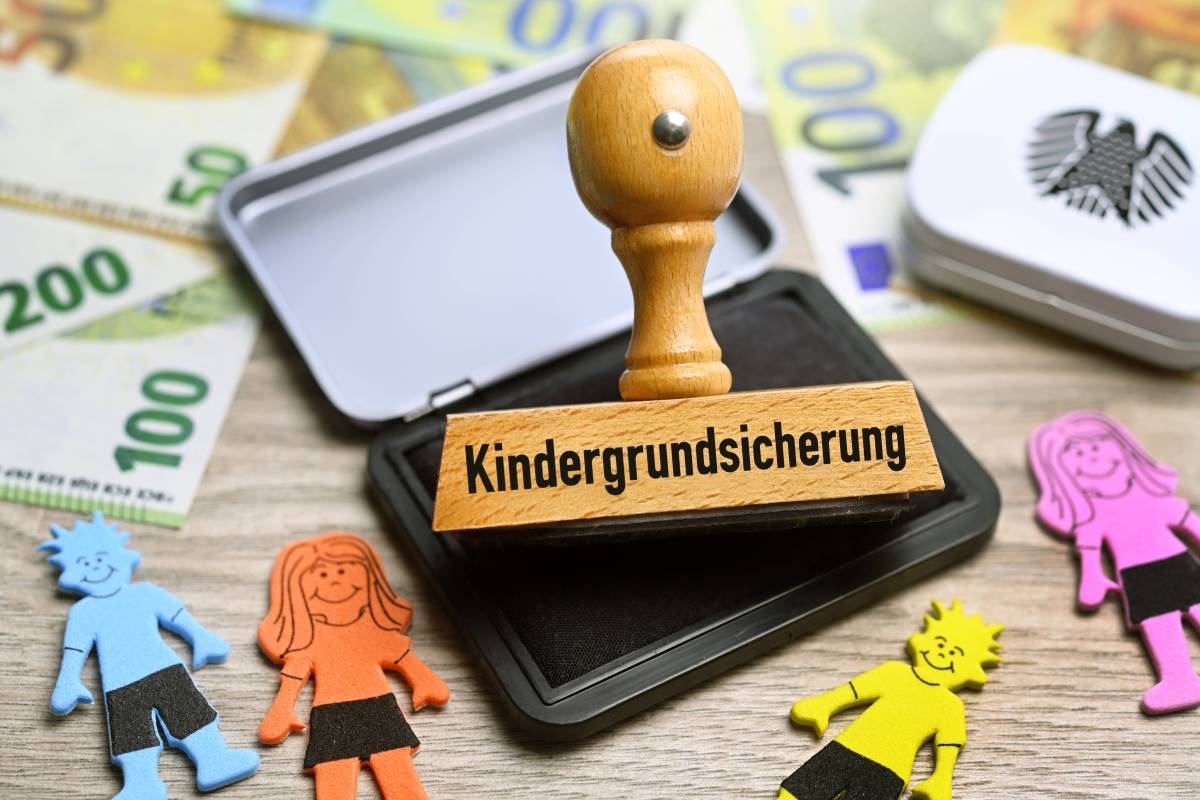 Stempel mit der Aufschrift "Kindergrundsicherung" liegt zwischen Geldscheinen und Kinderfiguren.