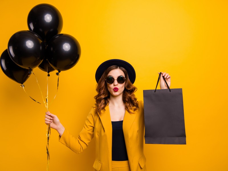 Eine Frau mit braunen Haaren und Sonnenbrille hält schwarze Luftballons und eine schwarze Tüte in der Hand.