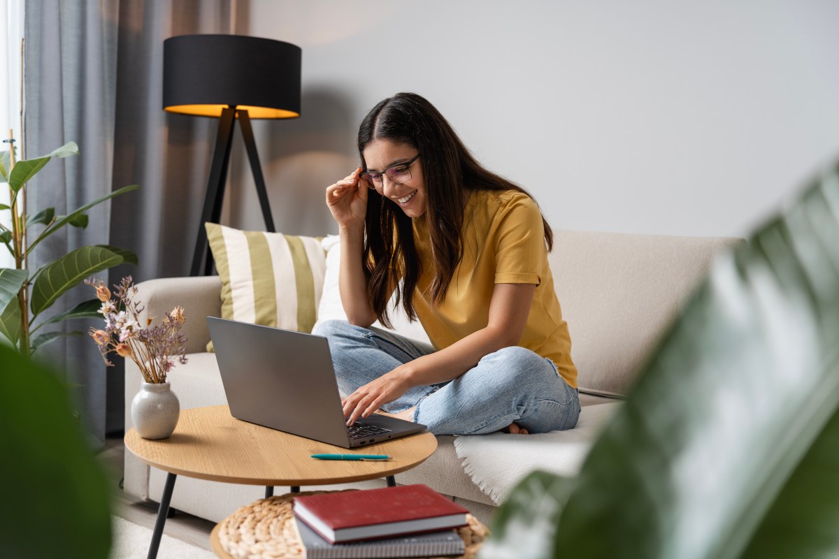 Eine Frau sitzt auf der Couch in ihrem Wohnzimmer und arbeitet am Laptop.