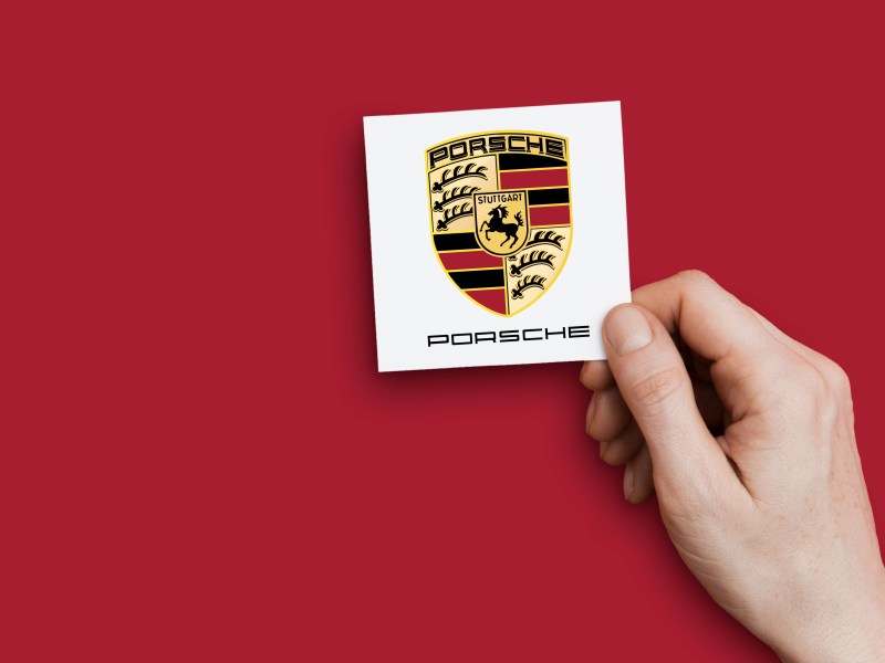 Eine Hand hÃ¤lt das Logo von Porsche vor eine rote Wand.