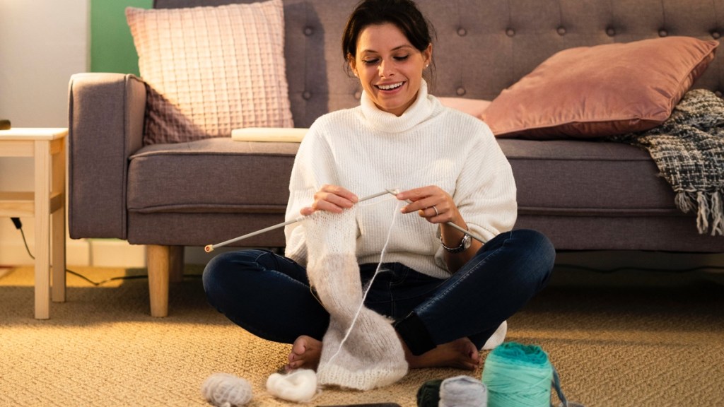 Eine strickende, lächelnde Frau sitzt vor dem Sofa.