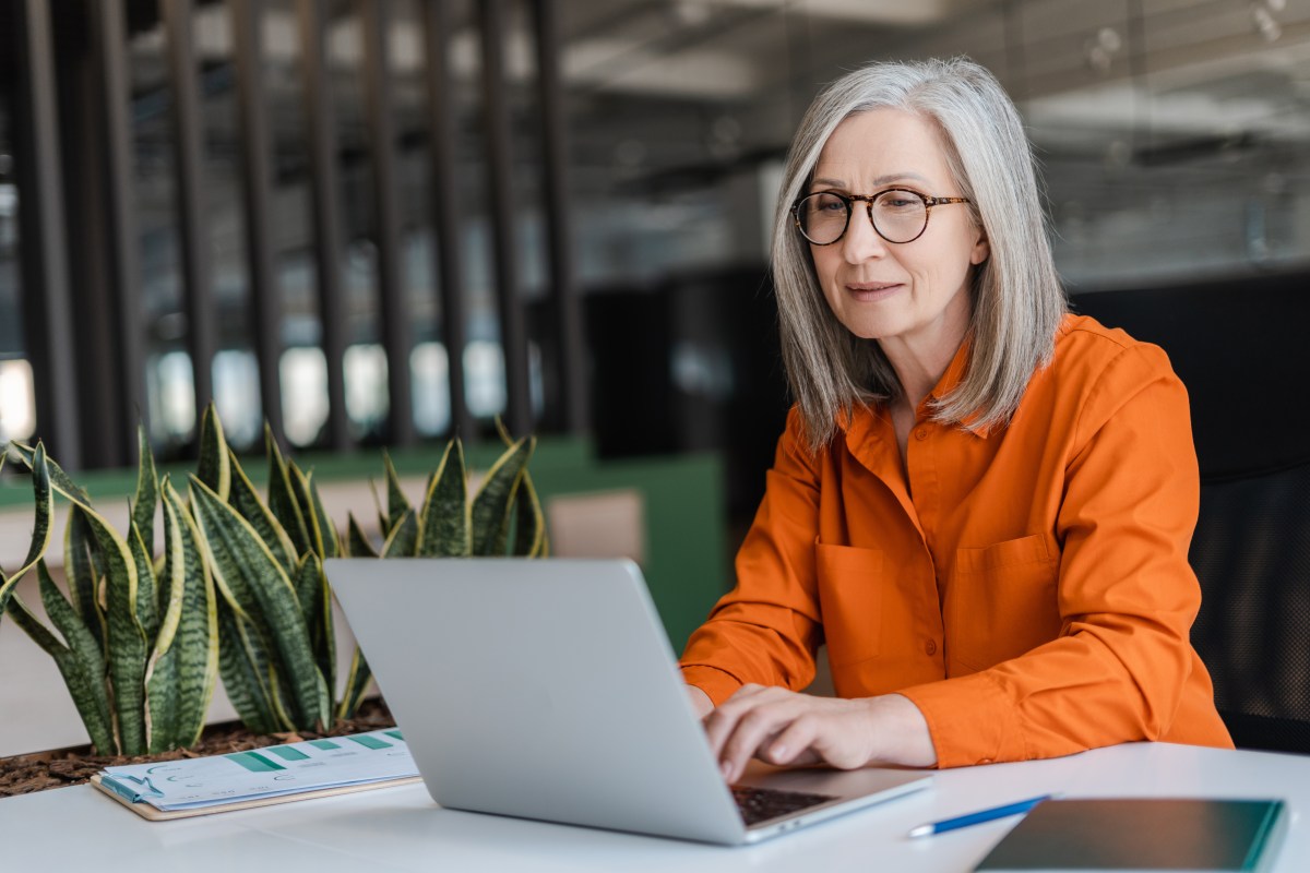 Eine ältere Dame mit grauen Haaren sitzt im Büro vor einem Laptop.