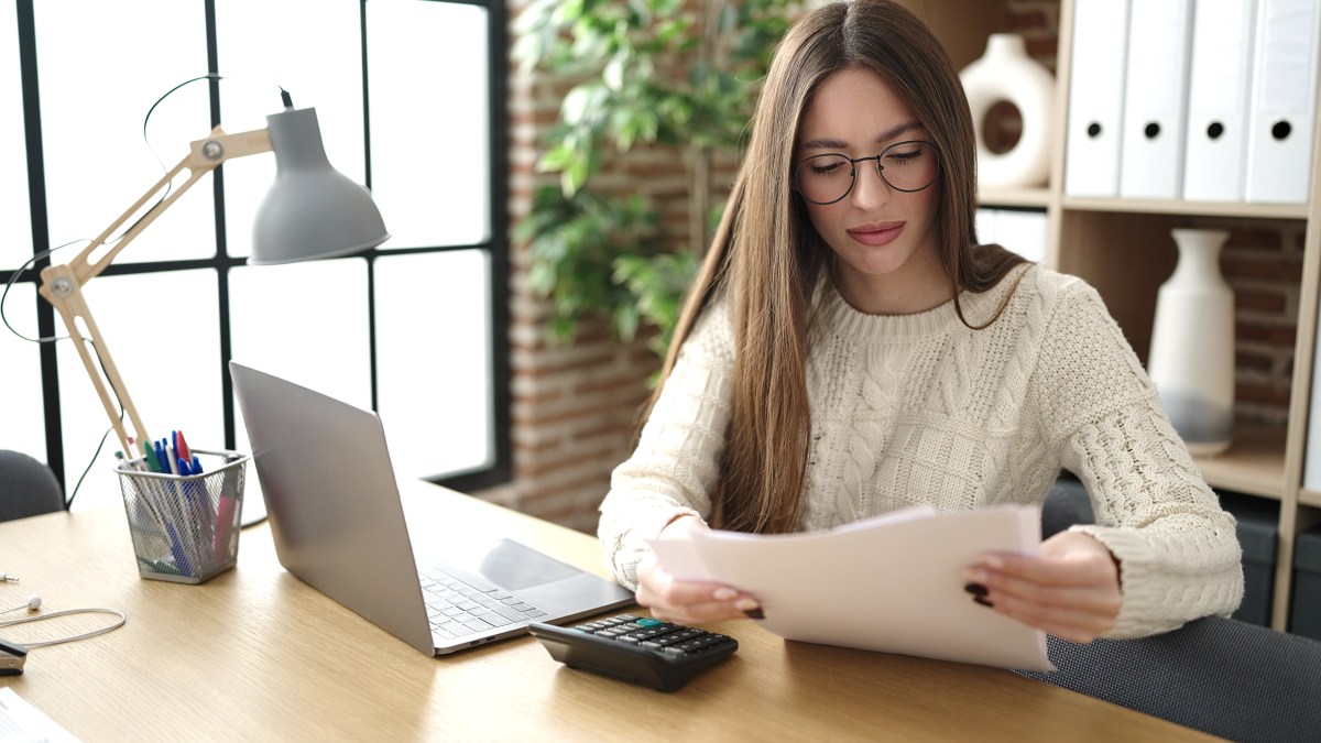 Eine Frau sitzt am Schreibtisch und schaut sich ein Dokument an. Neben ihr steht ein Laptop.