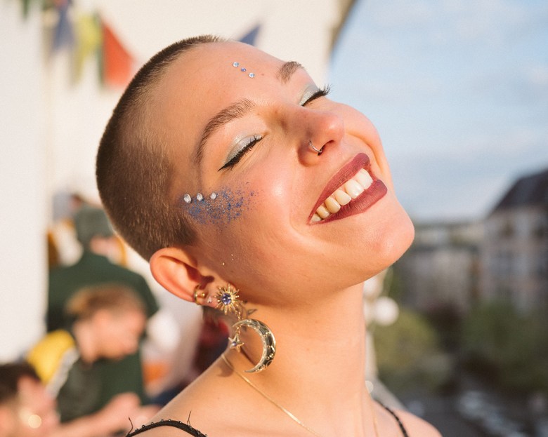 Frau mit rasierten Haaren lächelt und hat Glitzer Make-Up drauf