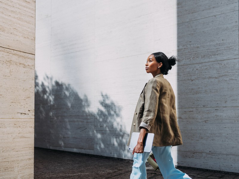 Eine selbstbewusste Geschäftsfrau in einem legeren Blazer und einer Hose geht mit einem Tablet in der Hand nach draußen.