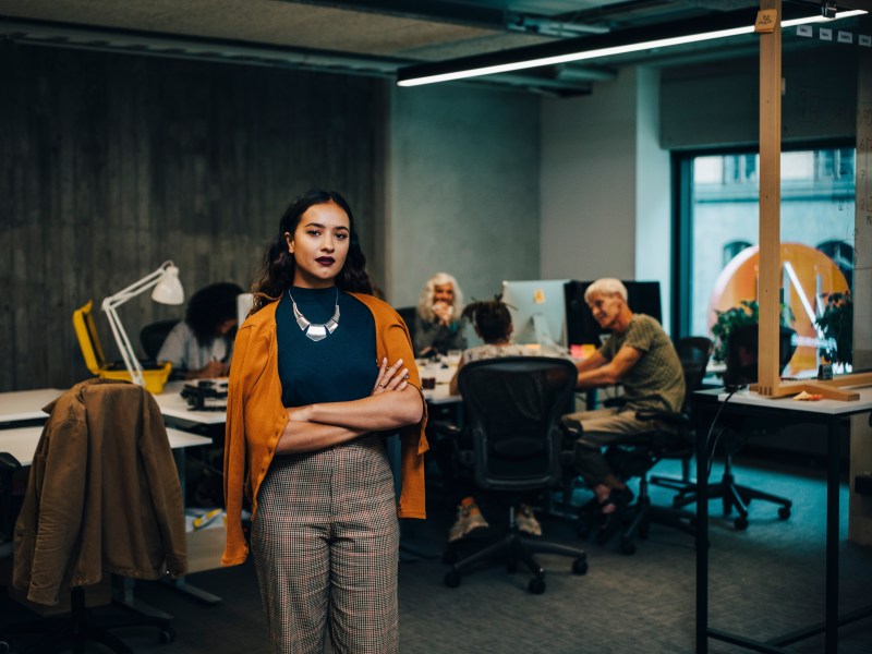Eine Frau mit dunklen haaren und dunklem Lippenstift steht mit verschränkten Armen in einem Großraumbüro.