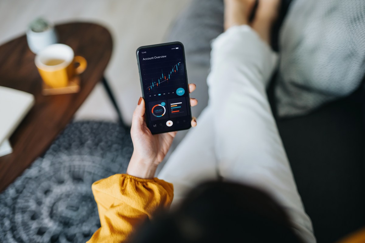 Blick von oben auf eine junge Frau, die Finanzen und Investitionen verwaltet und zu Hause Börsendaten auf dem Smartphone analysiert