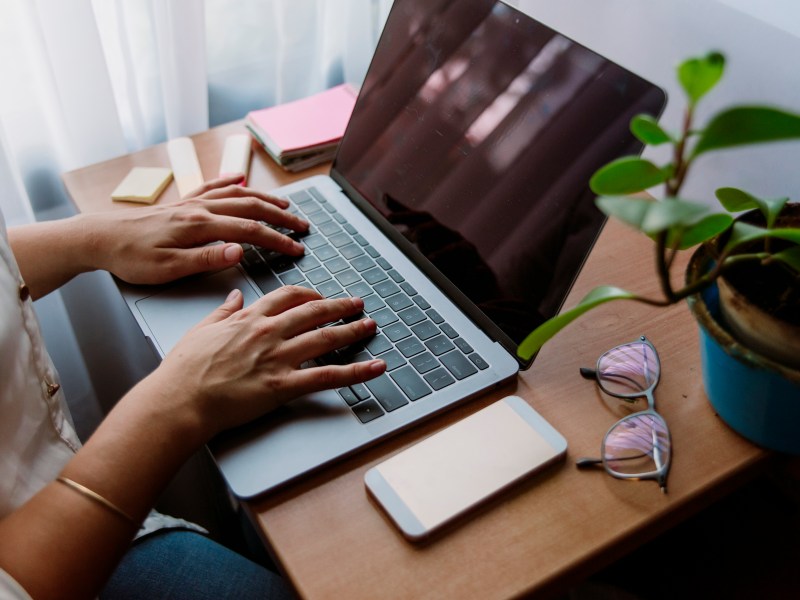 Frau schreibt am Laptop und arbeitet am Schreibtischarbeitsplatz mit Pflanzen, Mobiltelefon und Brille.