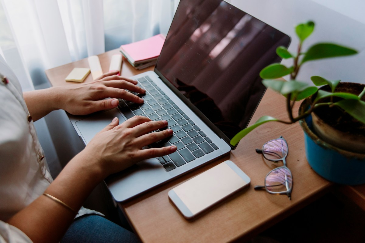 Frau schreibt am Laptop und arbeitet am Schreibtischarbeitsplatz mit Pflanzen, Mobiltelefon und Brille.