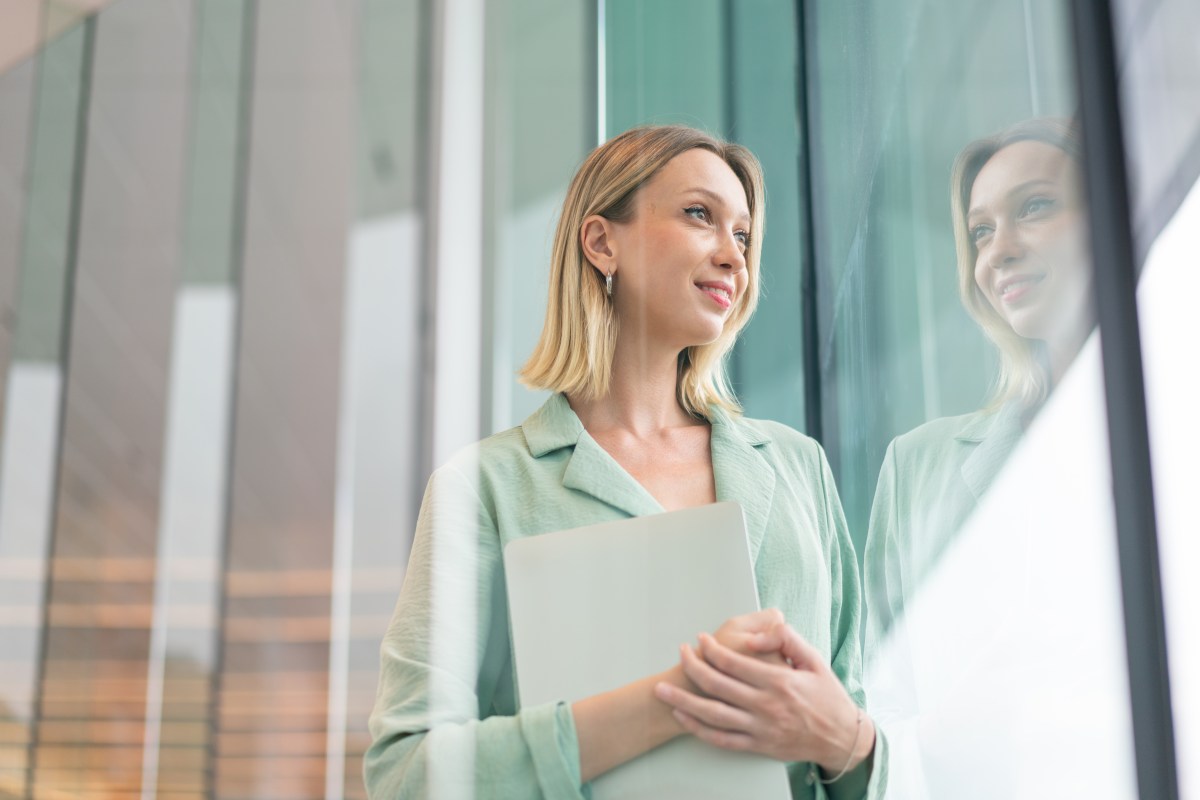 Blonde Frau im Mint-Blazer hält Laptop in der Nähe von Glasfenstern.