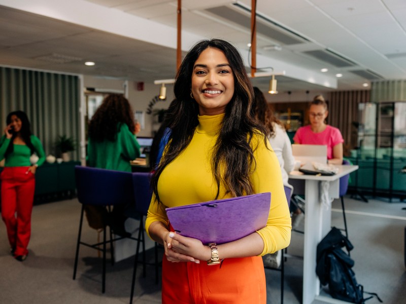 Eine Frau in einem gelber Oberteil steht im Büro und hält einen Ordner in der Hand.