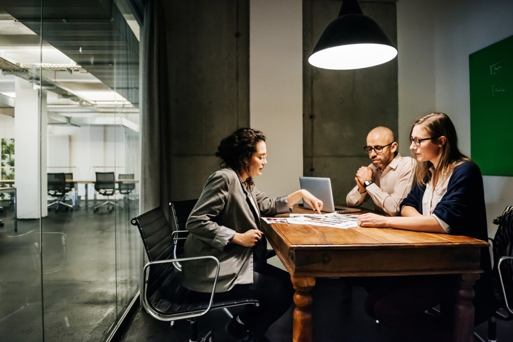 Drei Personen sitzen an einem alten Tisch in einem modernen Büro und halten ein Meeting ab.