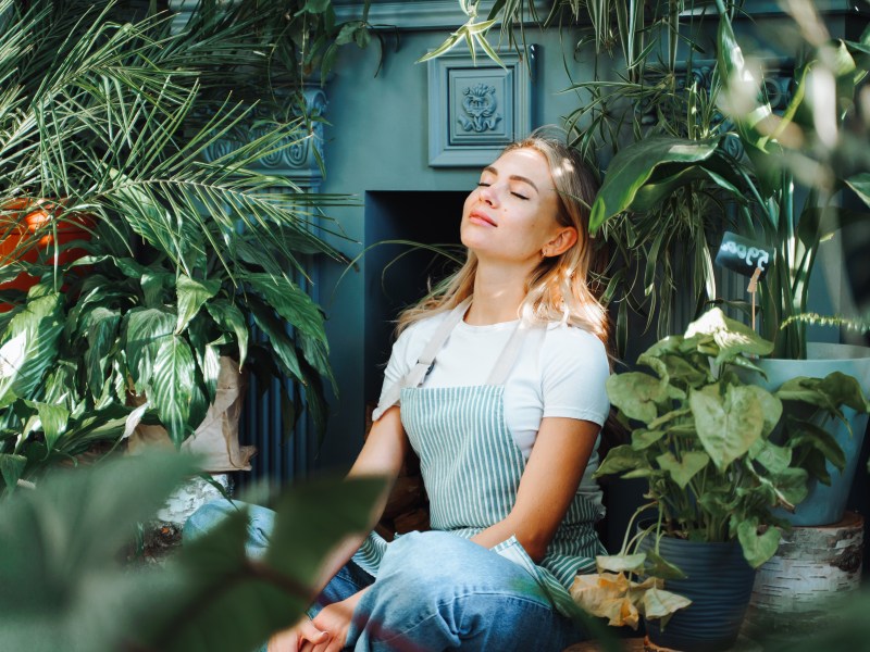 Pausen im Job: Entspannende und meditierende lächelnde Frau in der Nähe von grünen Pflanzen und Blumen.