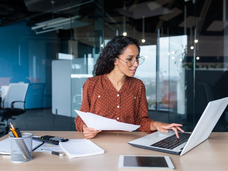Frau hält ein Dokument in der Hand und schaut etwas im Internet nach.