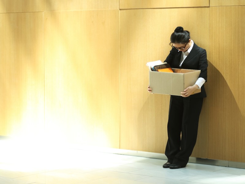 Eine Frau im Anzug lehnt an der Wand und schaut zu Boden. Sie trägt einen Karton mit Sachen.