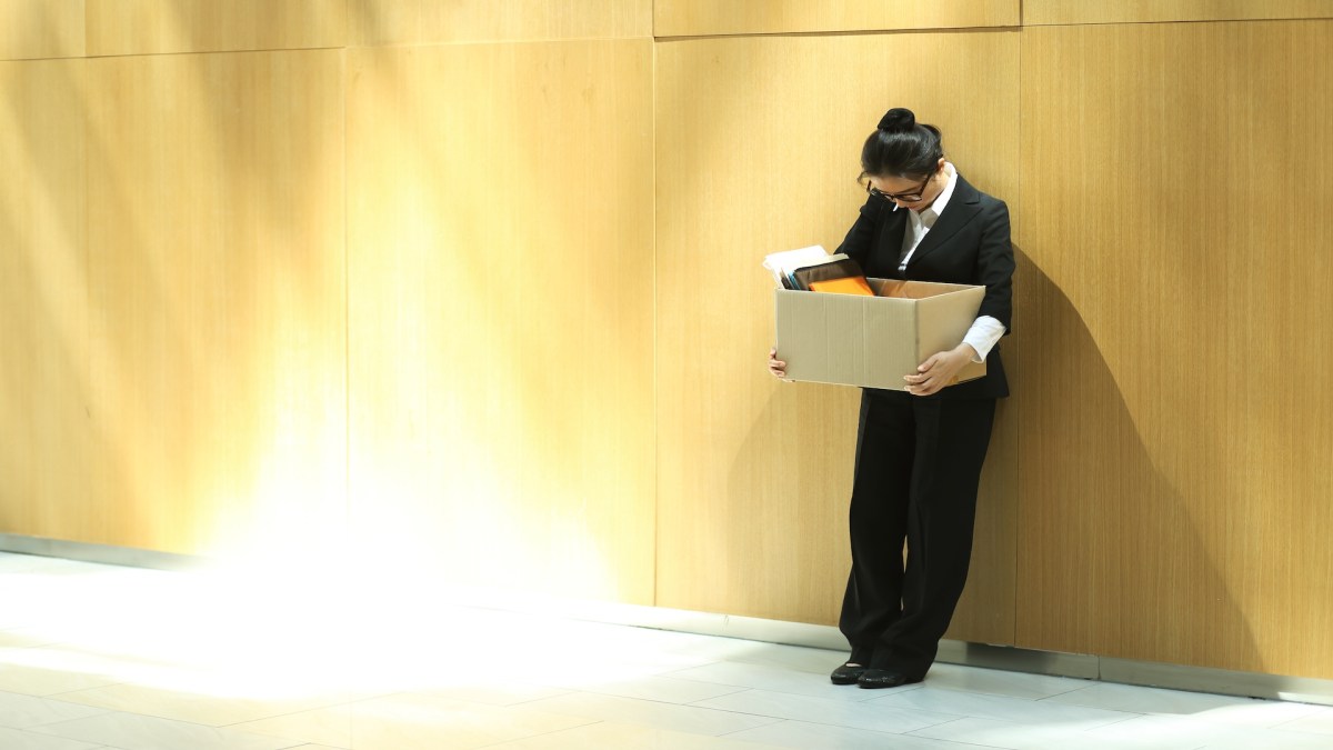 Eine Frau im Anzug lehnt an der Wand und schaut zu Boden. Sie trägt einen Karton mit Sachen.