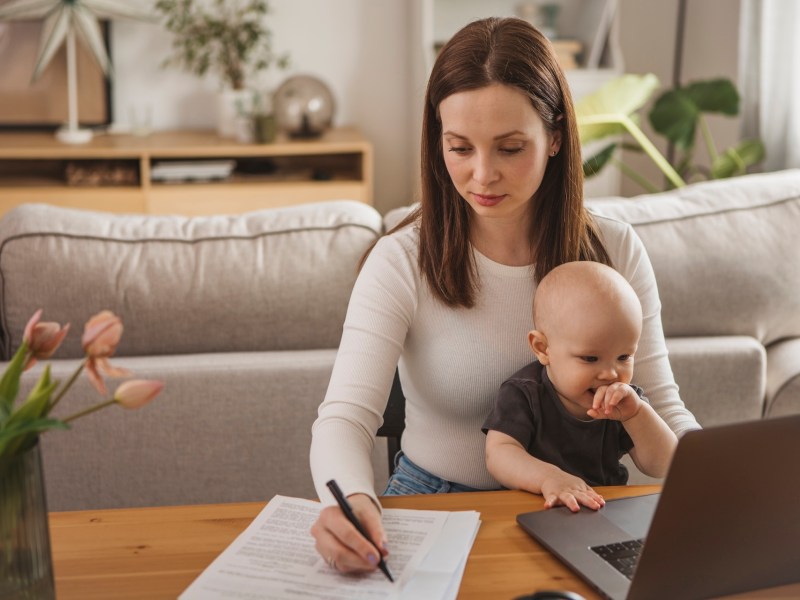 Junge Mutter mit Baby zu Hause, arbeitet am Laptop.