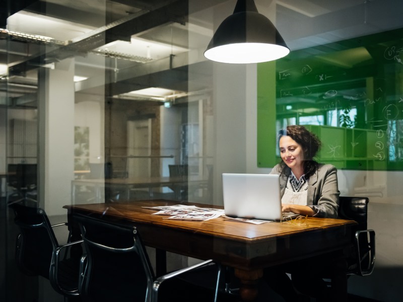 Eine Frau sitzt an einem Tisch in einem Büro und blickt zufrieden auf einen Laptop.