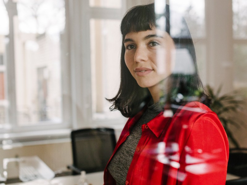 Eine Geschäftsfrau trägt ein rotes Hemd und steht im Büro hinter einer Glasfensterscheibe.