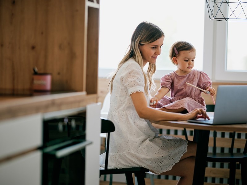 Eine Mutter sitzt in der Küche am Laptop. Daneben sitzt ein Kleinkind.