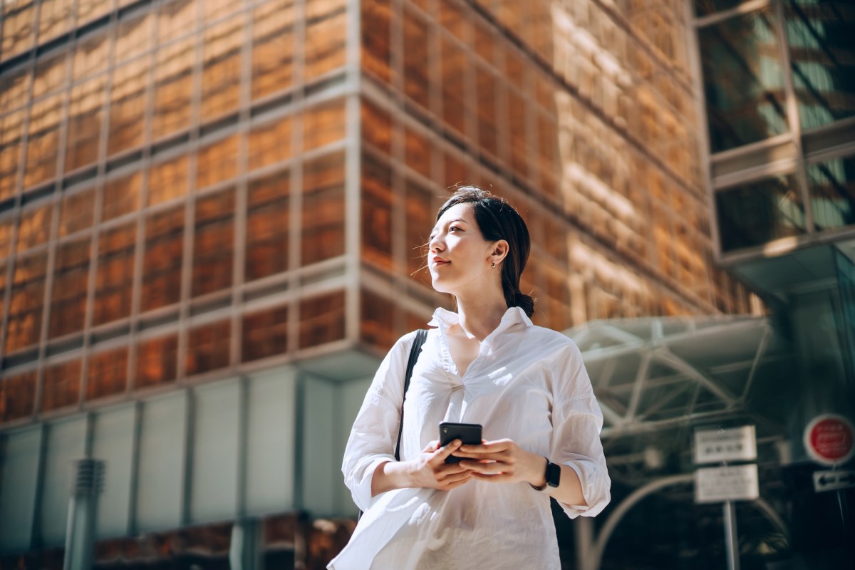 Eine junge Frau steht mit ihrem Handy vor einem Bürogebäude.