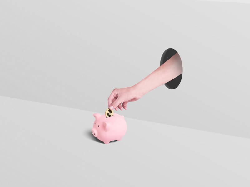 Eine Hand greift durch ein Loch und wirft eine Münze in ein Sparschwein.