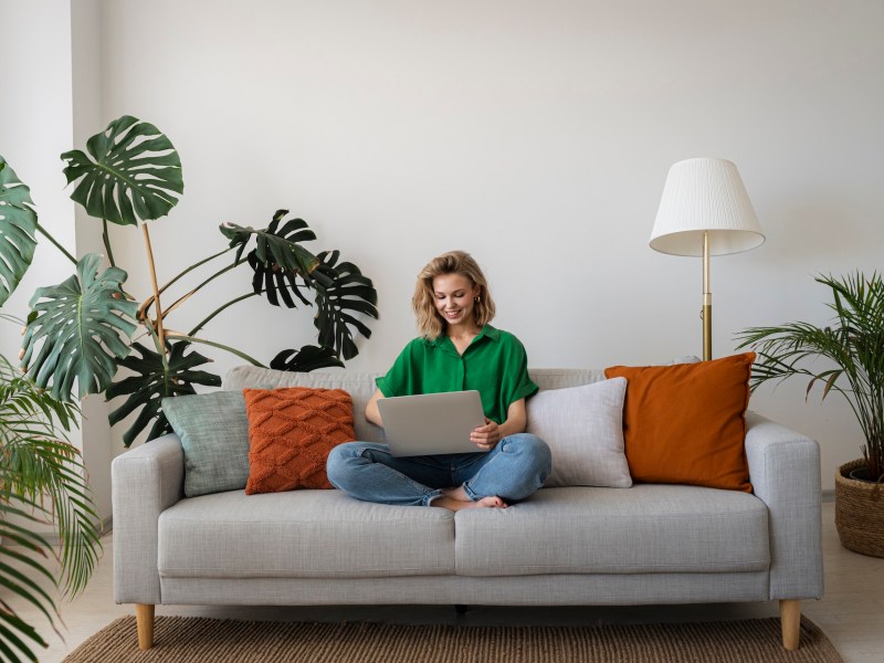 Eine junge Frau sitzt zu Hause auf dem Sofa und arbeitet am Laptop.