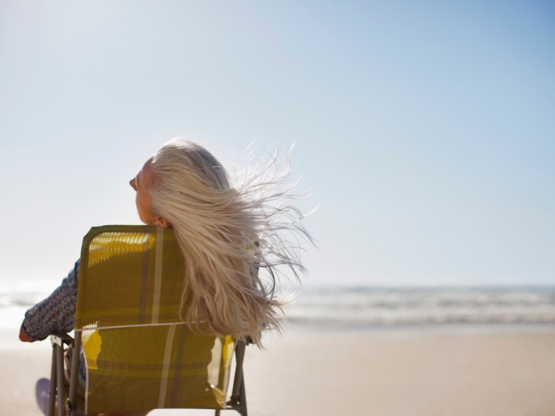 Eine Seniorin mit grauen Haaren sitzt am Meer in einem Liegestuhl.