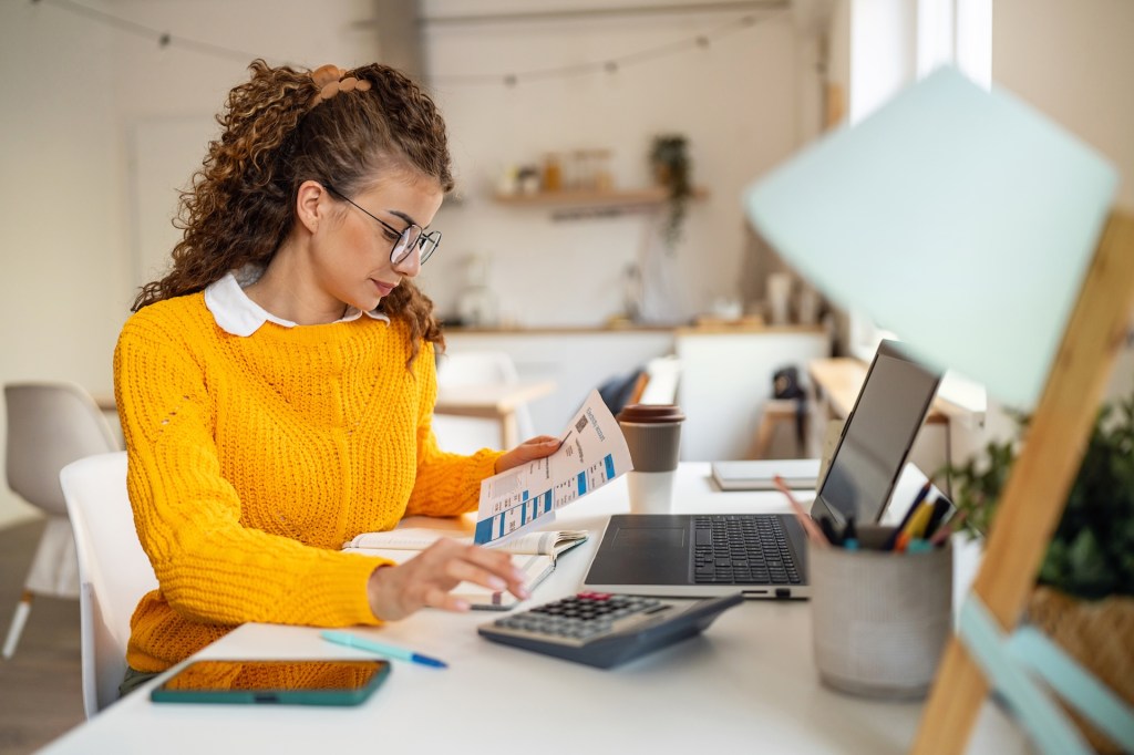 Eine junge Frau im gelben Pullover sitzt am Schreibtisch und schaut Steuerunterlagen durch.