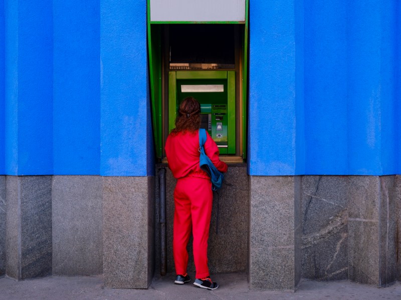 Frau in einem roten Bürooutfit hebt Geld an einem Automaten ab.