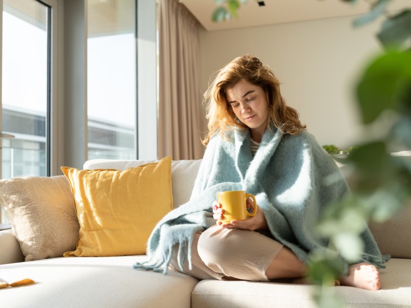 Junge Frau in Decke gehüllt macht eine Pause mit einer Tasse Tee im Wohnzimmer.