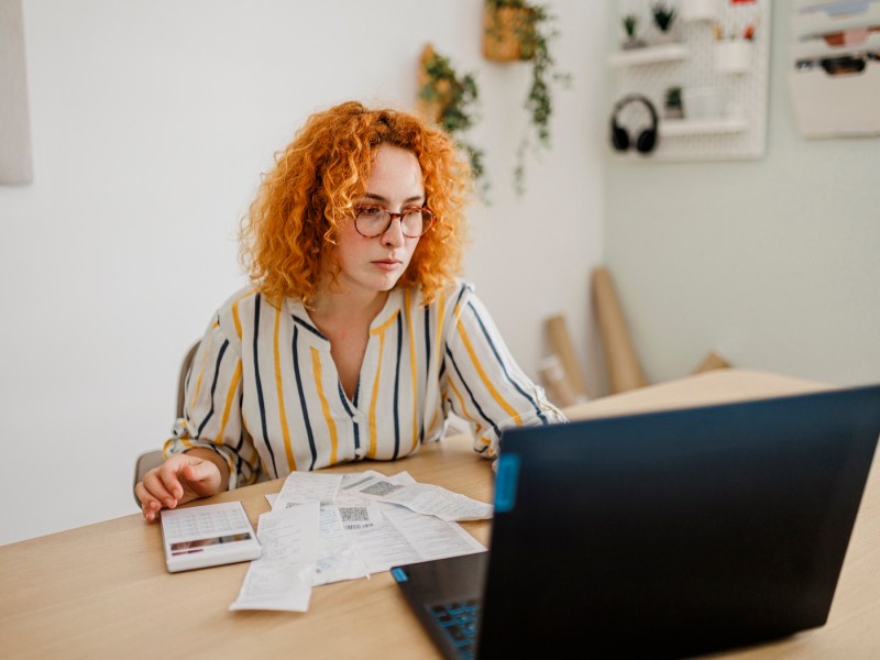 Eine junge Frau sitzt mit Belegen vor dem Laptop.