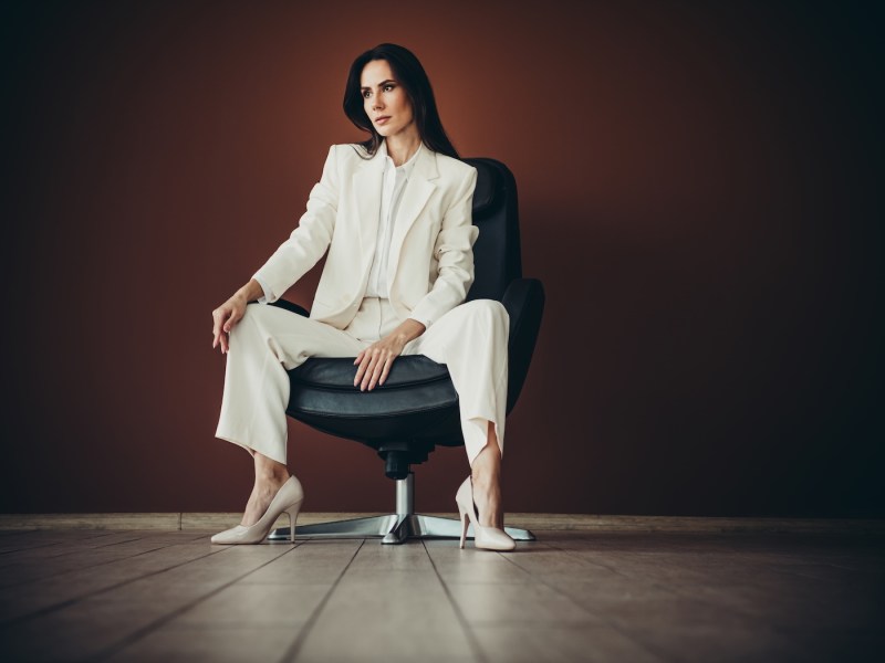 Eine berufstätige Frau im weißen Anzug sitzt selbstbewusst auf einem Stuhl.