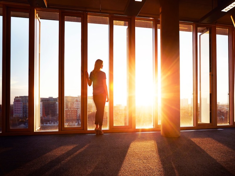 Eine Frau steht im Büro und schaut bei Sonnenuntergang aus dem Fenster.