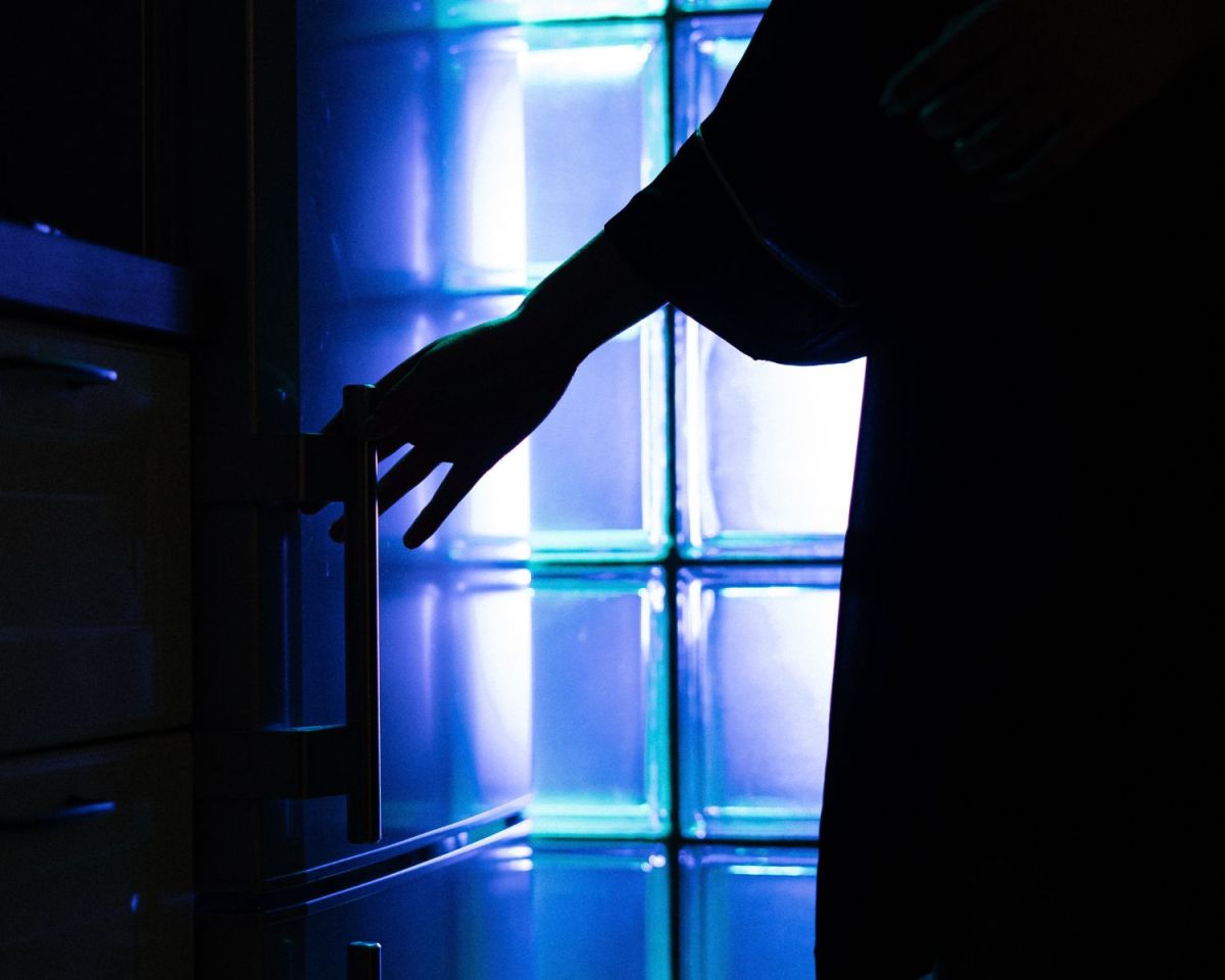 Arm öffnet einen Kühlschrank vor dem Hintergrund eines blau belichteten Fensters