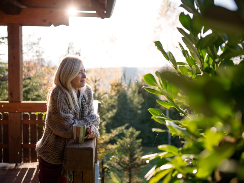 Porträt einer alten Frau, die neben einem Holzhaus steht.