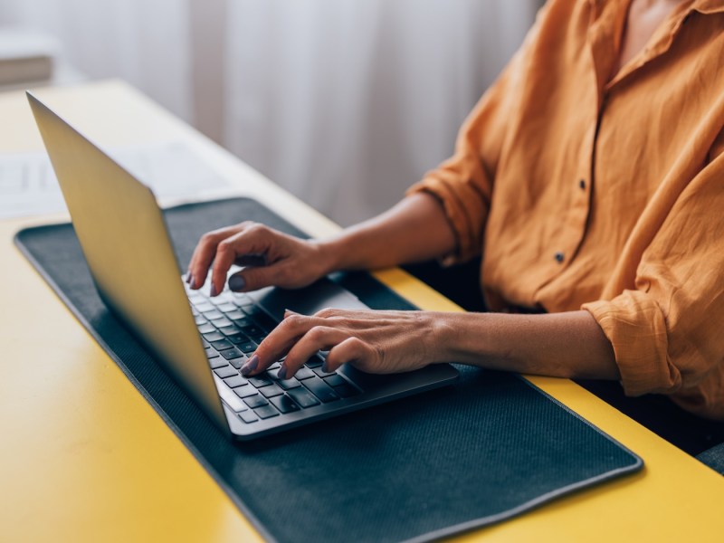 Nahaufnahme einer Frau in einem gelben Hemd, die an einem Arbeitsplatz mit Papierkram herumsitzt und auf einem Laptop tippt.