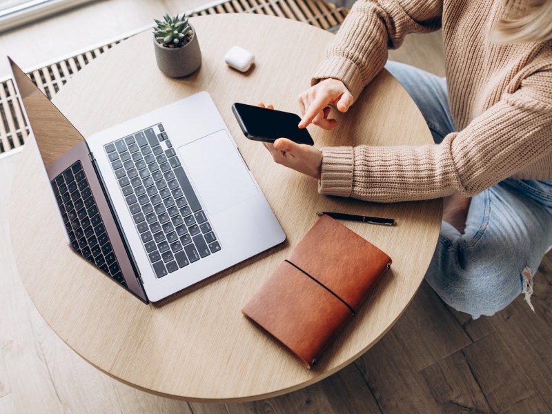 Draufsicht einer Frau mit Smartphone in der Hand und Laptop auf dem Tisch.