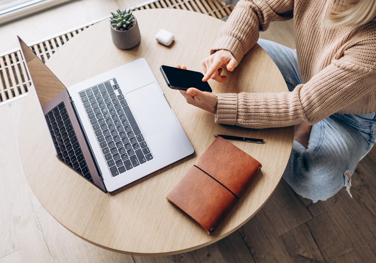 Draufsicht einer Frau mit Smartphone in der Hand und Laptop auf dem Tisch.
