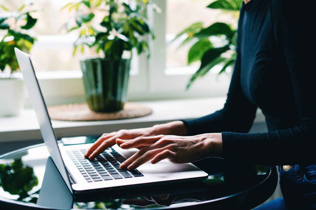 Frau tippt auf einem Laptop an einem Schreibtisch mit Fenstern und gr&uuml;nen Pflanzen.