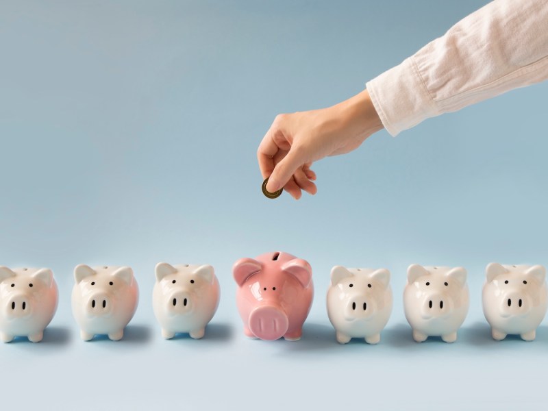 Hand wirft eine Münze in ein größeres rosa Sparschwein auf blauem Hintergrund.