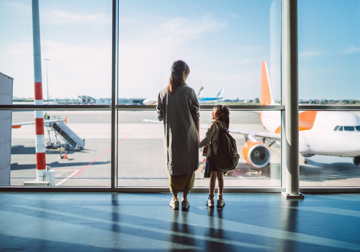 Rückansicht einer Mutter und ihrer kleinen Tochter, die am Fenster eines Flughafenterminals stehen und im Sonnenlicht die Flugzeuge auf dem Rollfeld beobachten.