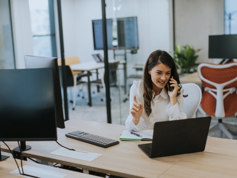 Geschäftsfrau, die im modernen Büro mit dem Mobiltelefon arbeitet, während sie am Laptop arbeitet.