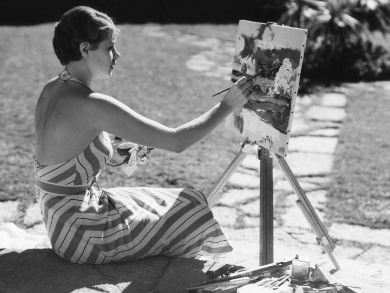 Frau sitzt vor einer Leinwand und malt in Schwarz-Weiß.