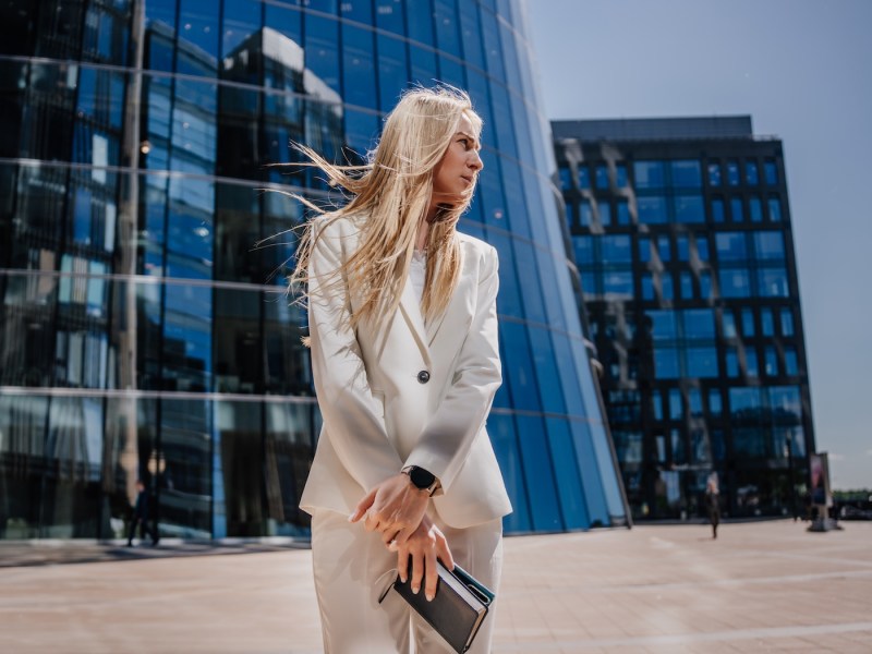 Junge blonde Frau im weißen Anzug steht draußen vor einem gläsernen Gebäude.